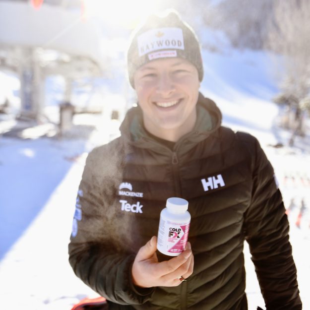 Brodie Seger standing on the snow in front of a chairlift, holding a container of ColdFX.